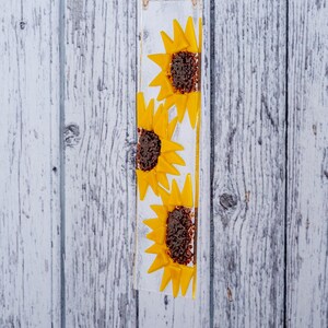 Puede incluir: Un panel de vidrio con tres girasoles amarillos con centros marrones. El panel est&aacute; colgado de una cuerda blanca.