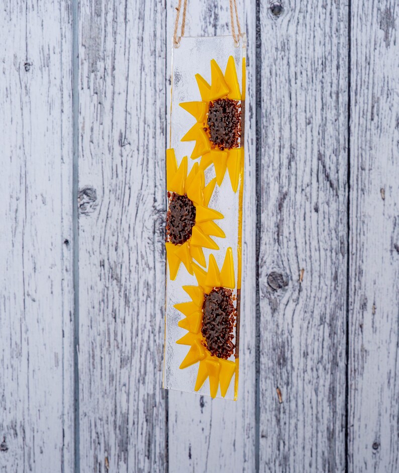 Puede incluir: Un panel de vidrio con tres girasoles amarillos con centros marrones. El panel est&aacute; colgado de una cuerda blanca.
