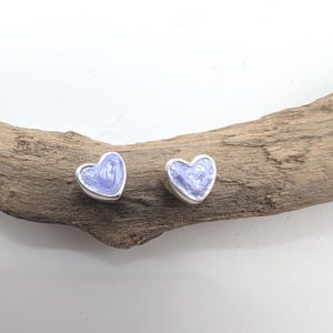 May include: A pair of heart-shaped stud earrings. Each earring features a silver border and a light purple enamel filling. The earrings are displayed on a piece of driftwood against a white background. These earrings are a charming accessory.