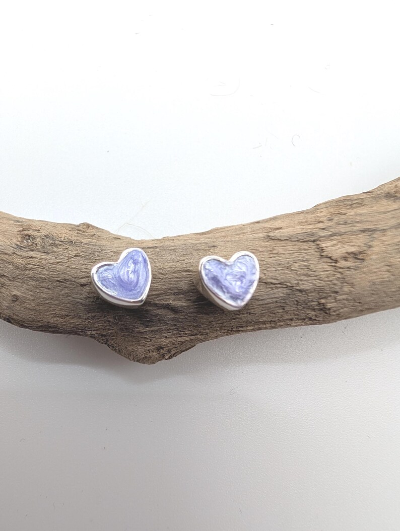 May include: A pair of heart-shaped stud earrings. Each earring features a silver border and a light purple enamel filling. The earrings are displayed on a piece of driftwood against a white background. These earrings are a charming accessory.