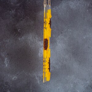 Puede incluir: Un adorno de vidrio transparente con un dise&ntilde;o de girasol amarillo. El girasol est&aacute; hecho de resina y tiene una superficie texturizada. El adorno est&aacute; colgado de una cuerda.