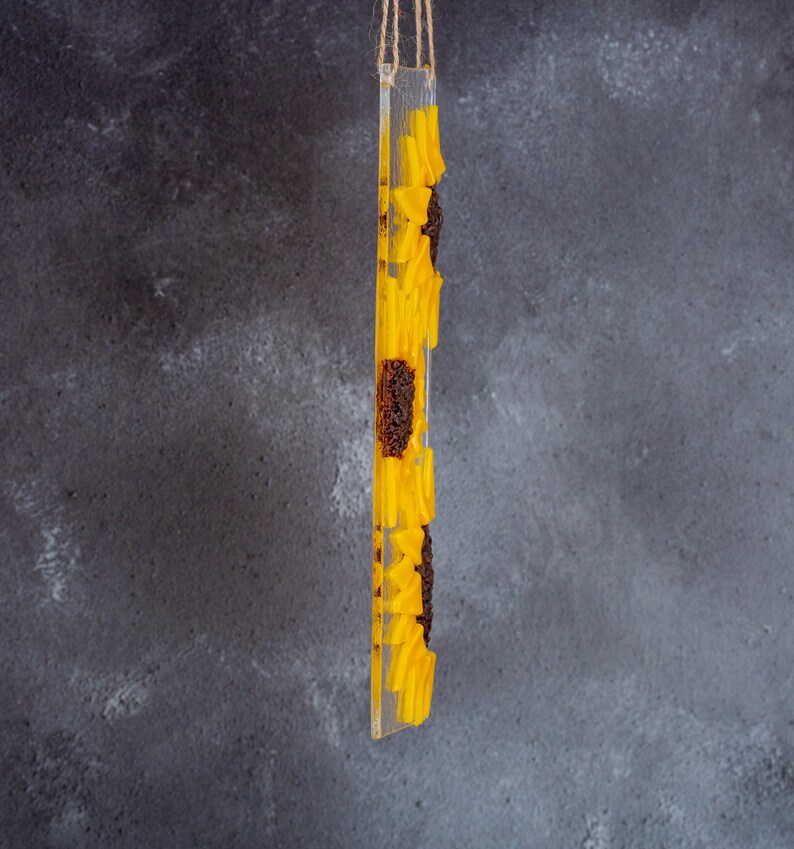 Puede incluir: Un adorno de vidrio transparente con un dise&ntilde;o de girasol amarillo. El girasol est&aacute; hecho de resina y tiene una superficie texturizada. El adorno est&aacute; colgado de una cuerda.