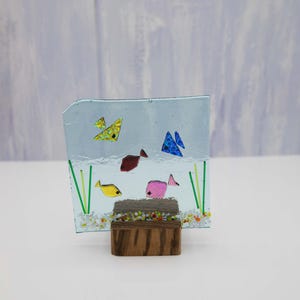 May include: A glass art piece depicting a fish tank with colorful fish and green plants on a wooden stand.