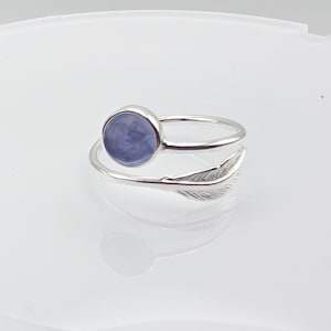 May include: A silver ring featuring a light purple, circular stone and a feather design. The ring has a split shank, with the feather detail wrapping around the finger. The stone is set in a bezel setting.