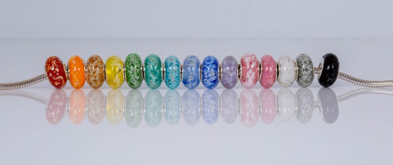 Op de afbeelding: Een regenboog van kleurrijke glazen kralen met zilveren accenten op een zilveren ketting. De kralen zijn bespikkeld met witte vlekken.