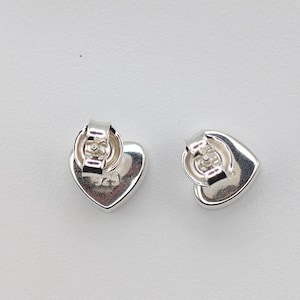 May include: Two silver heart-shaped stud earrings. The earrings have a polished finish and a post back. The back of each earring is stamped with the number "925". The earrings are displayed on a white surface.