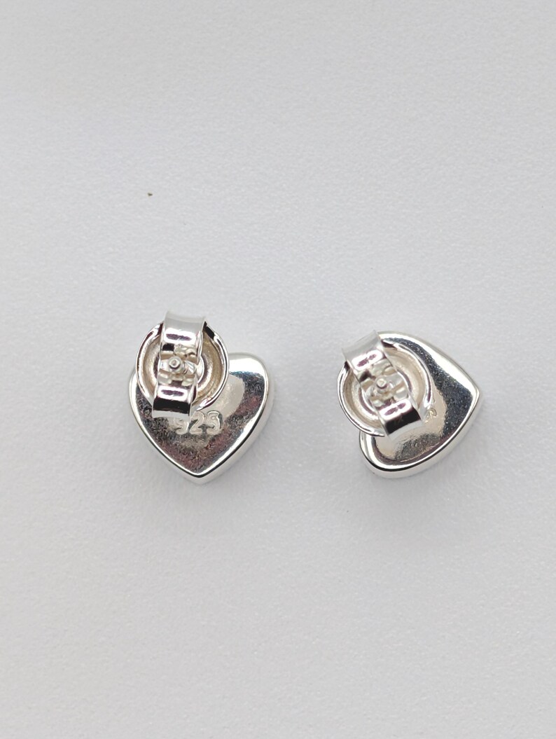 May include: Two silver heart-shaped stud earrings. The earrings have a polished finish and a post back. The back of each earring is stamped with the number "925". The earrings are displayed on a white surface.
