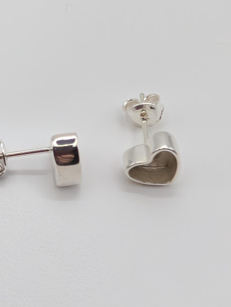May include: Two silver heart-shaped stud earrings. The earrings are made of a shiny, reflective metal. The heart shape is simple and smooth, with a slightly rounded form. The posts and backs are also silver.