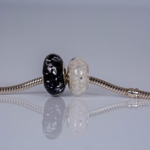 Op de afbeelding: Een zilveren armband met twee glazen kralen, een zwarte met witte stippen en een witte met zwarte stippen.