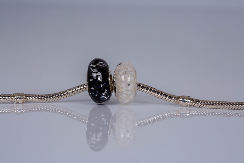 Op de afbeelding: Een zilveren armband met twee glazen kralen, een zwarte met witte stippen en een witte met zwarte stippen.