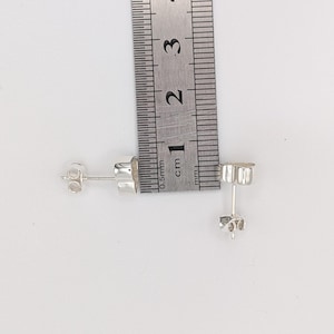 May include: A pair of small, silver stud earrings with butterfly backs, next to a ruler. The earrings are approximately 0.5 cm in diameter. The ruler shows measurements in centimeters.