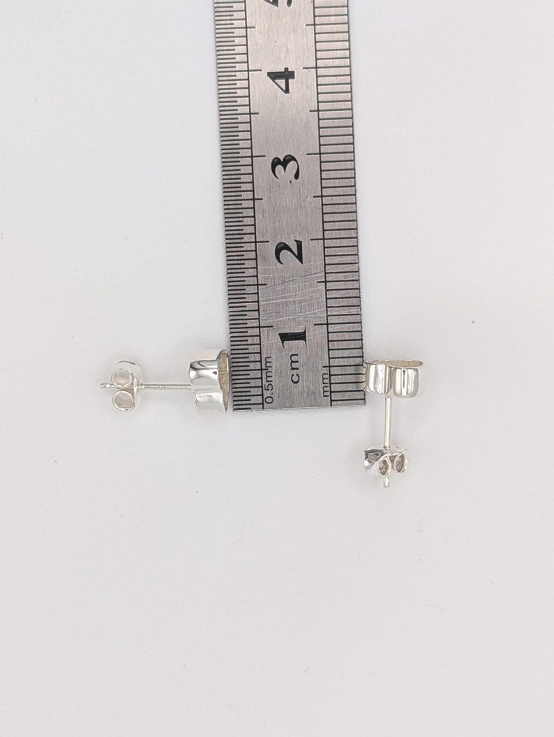 May include: A pair of small, silver stud earrings with butterfly backs, next to a ruler. The earrings are approximately 0.5 cm in diameter. The ruler shows measurements in centimeters.