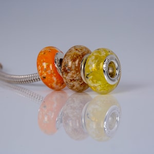 Op de afbeelding: Drie glazen kralen met zilveren accenten. De kralen zijn oranje, bruin en geel met witte vlekken. De kralen zijn geregen aan een zilveren ketting.