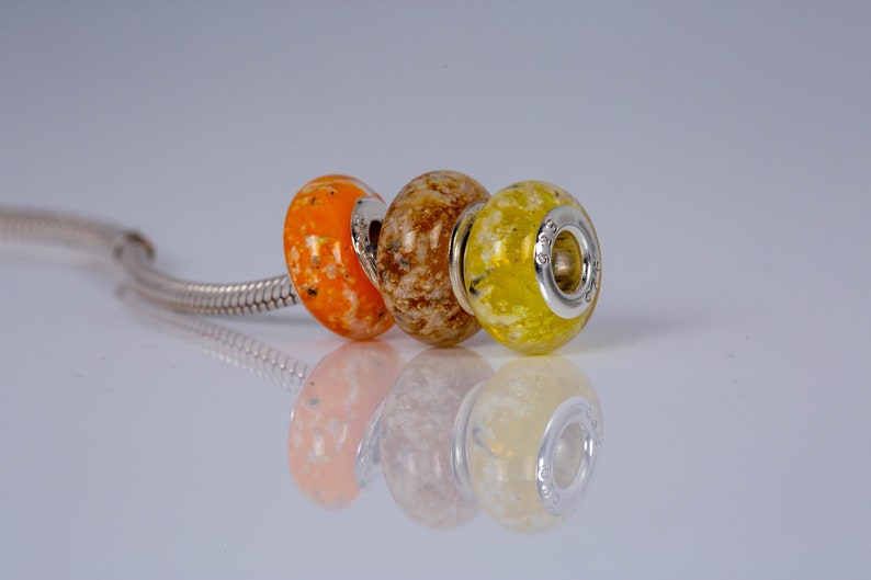 Op de afbeelding: Drie glazen kralen met zilveren accenten. De kralen zijn oranje, bruin en geel met witte vlekken. De kralen zijn geregen aan een zilveren ketting.