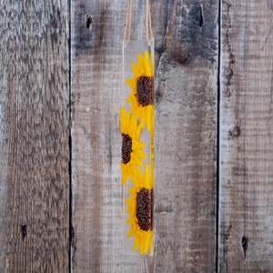 Puede incluir: Una suspensi&oacute;n de pared rectangular de resina transparente con tres girasoles amarillos prensados en su interior. Los girasoles est&aacute;n dispuestos verticalmente, con el girasol superior orientado hacia la derecha, el girasol del medio orientado hacia la izquierda y el girasol inferior orientado hacia la derecha.