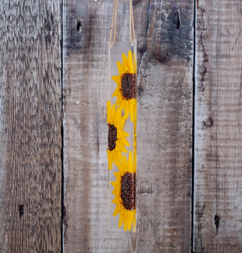 Puede incluir: Una suspensi&oacute;n de pared rectangular de resina transparente con tres girasoles amarillos prensados en su interior. Los girasoles est&aacute;n dispuestos verticalmente, con el girasol superior orientado hacia la derecha, el girasol del medio orientado hacia la izquierda y el girasol inferior orientado hacia la derecha.