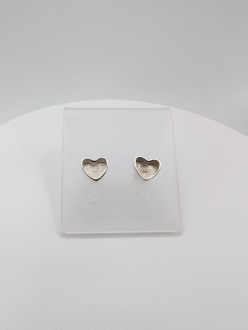 May include: A pair of silver heart-shaped stud earrings. The earrings are displayed on a clear plastic stand against a white background. The heart shapes have a slightly textured surface.