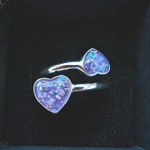 Könnte beinhalten: Ein silberfarbener Bypass-Ring mit zwei herzförmigen Fassungen. Jedes Herz enthält eine violette Harzeinlage, die mit irisierenden, mehrfarbigen Glitzerpartikeln gefüllt ist, welche das Licht einfangen und einen schimmernden Effekt erzeugen.