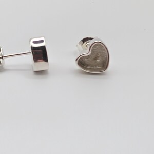May include: Silver-toned heart-shaped stud earrings. The earrings have a simple, minimalist design with a heart-shaped setting. The posts are visible, and the earrings are set against a white background.