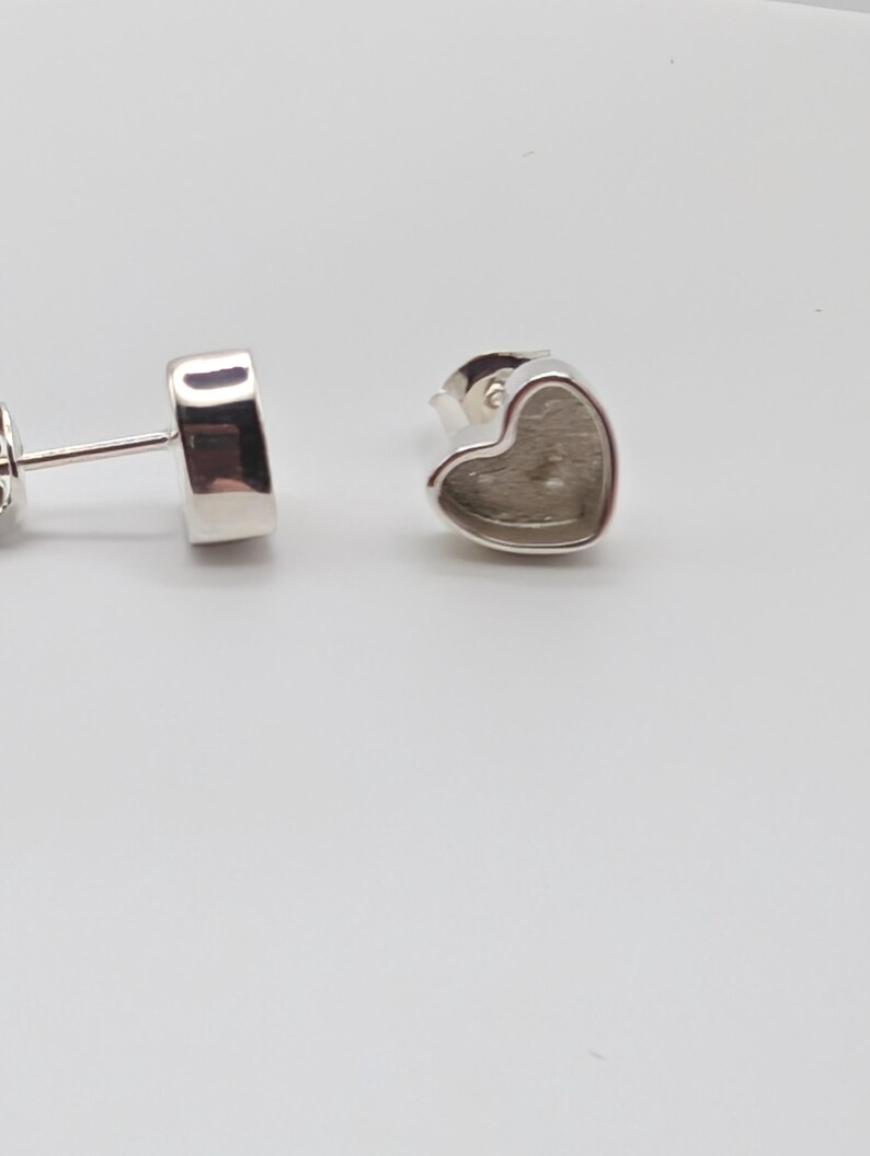 May include: Silver-toned heart-shaped stud earrings. The earrings have a simple, minimalist design with a heart-shaped setting. The posts are visible, and the earrings are set against a white background.