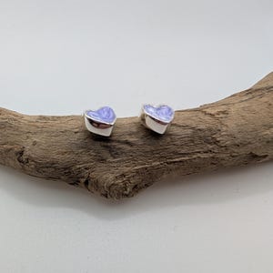 May include: Two silver heart-shaped stud earrings with a light purple enamel finish. The earrings are displayed on a piece of driftwood against a white background. These earrings are a charming accessory for everyday wear.