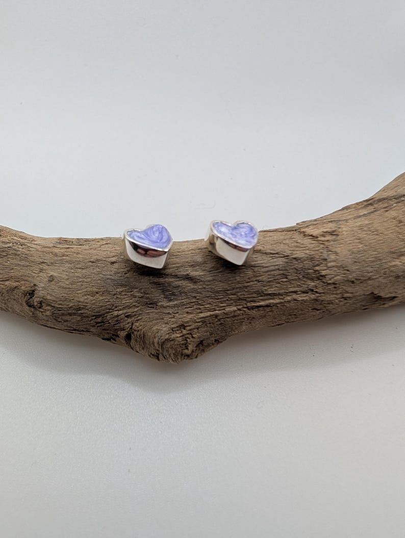 May include: Two silver heart-shaped stud earrings with a light purple enamel finish. The earrings are displayed on a piece of driftwood against a white background. These earrings are a charming accessory for everyday wear.