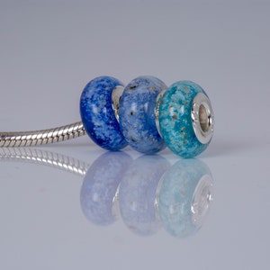 Op de afbeelding: Drie glazen kralen in tinten blauw, lichtblauw en turquoise, met zilveren accenten, op een zilveren slangenkettingarmband.
