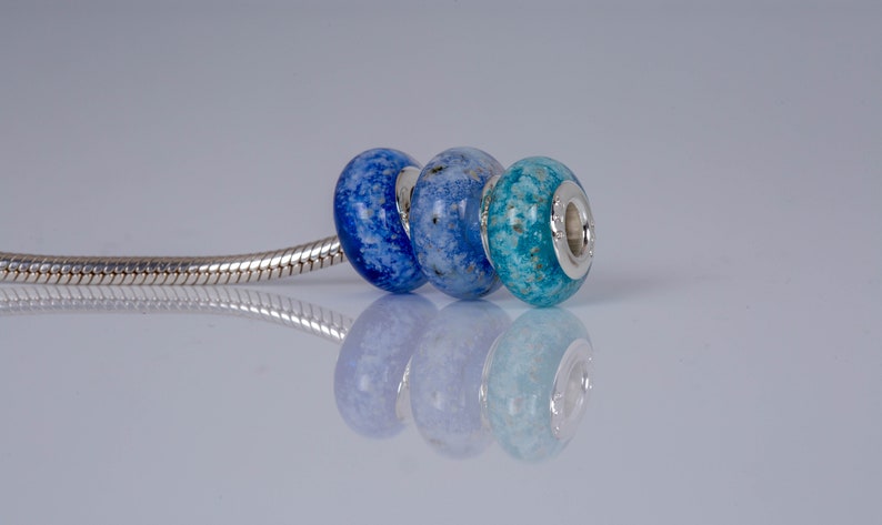 Op de afbeelding: Drie glazen kralen in tinten blauw, lichtblauw en turquoise, met zilveren accenten, op een zilveren slangenkettingarmband.