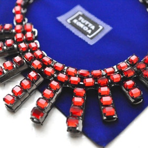 Può includere: Una collana di cristalli rossi con cornice in metallo argentato. La collana è su uno sfondo blu con il testo "Tutta la Moda" in un riquadro bianco.