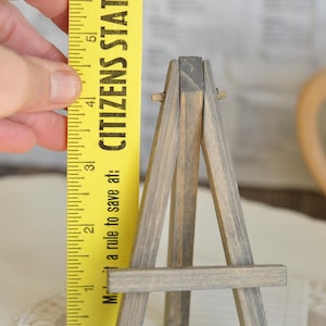May include: A small wooden easel, painted gray, is standing on a white surface. A yellow ruler with the words "Citizens State" and "Make a rule to save at:" is next to the easel.