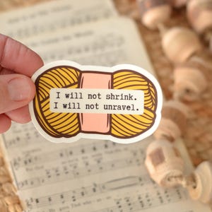 Puede incluir: Una pegatina con un diseño de ovillo de lana amarillo y el texto "I will not shrink. I will not unravel." La pegatina está sujeta por una mano, con partituras musicales en el fondo. También se ven cuentas de madera decorativas.