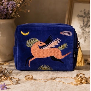 May include: A rectangular royal blue velvet pouch featuring an embroidered peach-colored winged horse, a yellow crescent moon, and a shooting star. The zipper pull is adorned with a yellow tassel, and a small black tag with white text is attached.