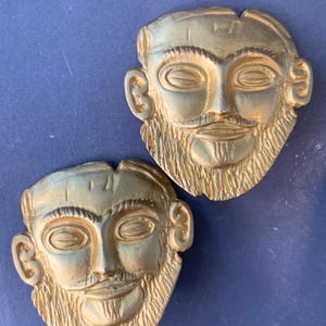 Puede incluir: Dos máscaras faciales de metal de color dorado con detalles precisos. Las máscaras tienen una expresión sonriente y barbuda.