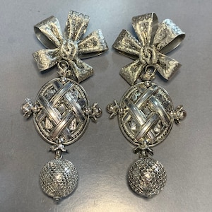 Puede incluir: Un par de pendientes colgantes de tono plateado con un lazo en la parte superior y un círculo tejido con una bola en la parte inferior.