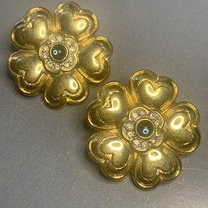 Op de afbeelding: Een paar gouden bloemvormige oorbellen met een zwarte steen en heldere strass steentjes in het midden.