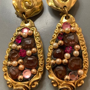 Puede incluir: Un par de pendientes colgantes de tono dorado con un diseño en forma de lágrima. Los pendientes están decorados con cuentas y pedrería marrones, rosas y doradas.