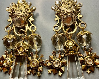 Op de afbeelding: Een paar goudkleurige hangende oorbellen met een kroonvormige bovenkant met heldere strass-steentjes, twee gefacetteerde heldere glaskralen en een centrale heldere kristallen hanger, afgewerkt met gedetailleerd metaalwerk.