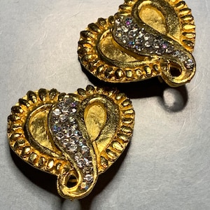 Pode incluir: Um par de brincos em forma de coração dourados com um design de espiral e strass transparentes.