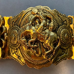 Puede incluir: Una hebilla de cinturón dorada con un diseño intrincado que presenta un jinete a caballo. La hebilla es adornada y detallada, con un diseño en relieve del jinete y el caballo.