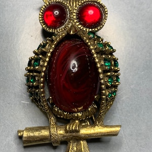 Op de afbeelding: Een gouden uilenbroche met rode en groene strass steentjes. De uil heeft een grote rode cabochon in het midden van zijn lichaam.