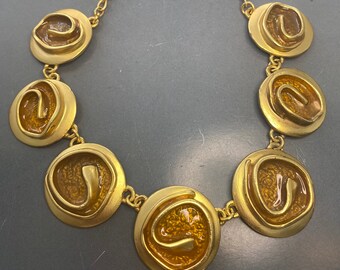 May include: A gold necklace with a chain and seven round pendants. Each pendant has a raised, textured, swirl design in a burnt orange color.