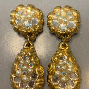 May include: A pair of gold tone dangle earrings with a teardrop shape. The earrings are adorned with clear and iridescent rhinestones and white pearls.