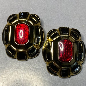 Puede incluir: Un par de pendientes de tono dorado con esmalte negro y piedras de vidrio rojo. Los pendientes tienen forma de flor con una piedra central redonda.