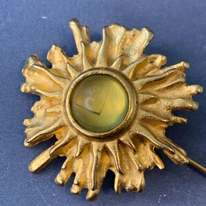 May include: A gold-toned brooch in the shape of a sun with a yellow glass center. The sun has a textured, organic design.
