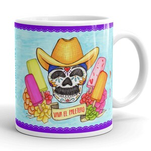 May include: White ceramic mug with a colorful illustration of a sugar skull wearing a yellow cowboy hat and surrounded by popsicles. The text "Viva el Paletero" is printed on a banner below the skull.