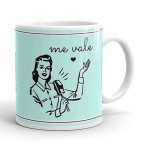 May include: White ceramic mug with a light blue background and a black and white illustration of a woman holding a microphone. The text "me vale" is printed in black on the mug.