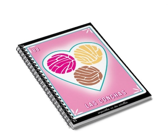 May include: A pink and black spiral notebook with a design of three colorful pastries in a heart shape. The text "LAS CONCHAS" is printed on the notebook, along with the words "serpientes vs dos estrellas".