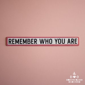May include: A pink and white rectangular sign with the text "REMEMBER WHO YOU ARE" in black lettering.
