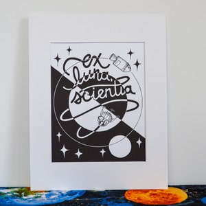 May include: Black and white illustration of a rocket ship and a moon with the text "Ex Luna, Scientia" in a stylized font.