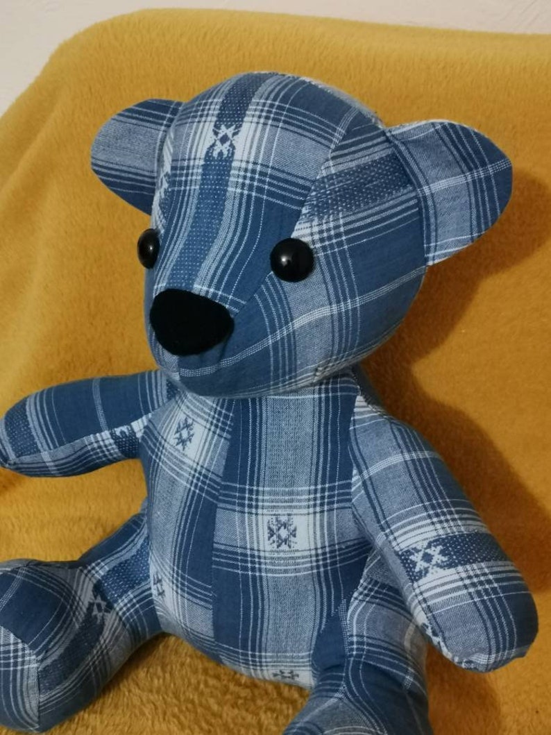 Pu&ograve; includere: Un orsacchiotto di peluche blu e bianco a quadri con occhi neri a bottone e un naso nero. L'orsacchiotto &egrave; seduto su una coperta gialla.